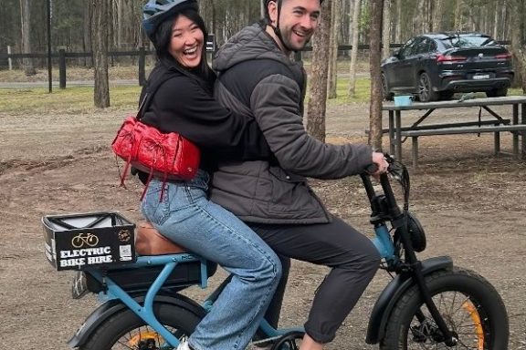 Two people wearing helmets on an electric bike, smiling, in a wooded area.